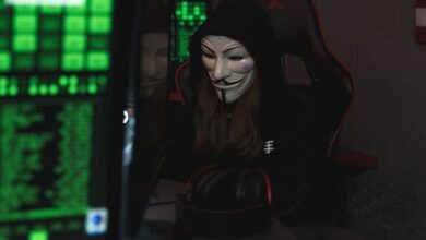 A mysterious hacker wearing a Guy Fawkes mask and black hoodie in a dimly lit room focused on computer screens.